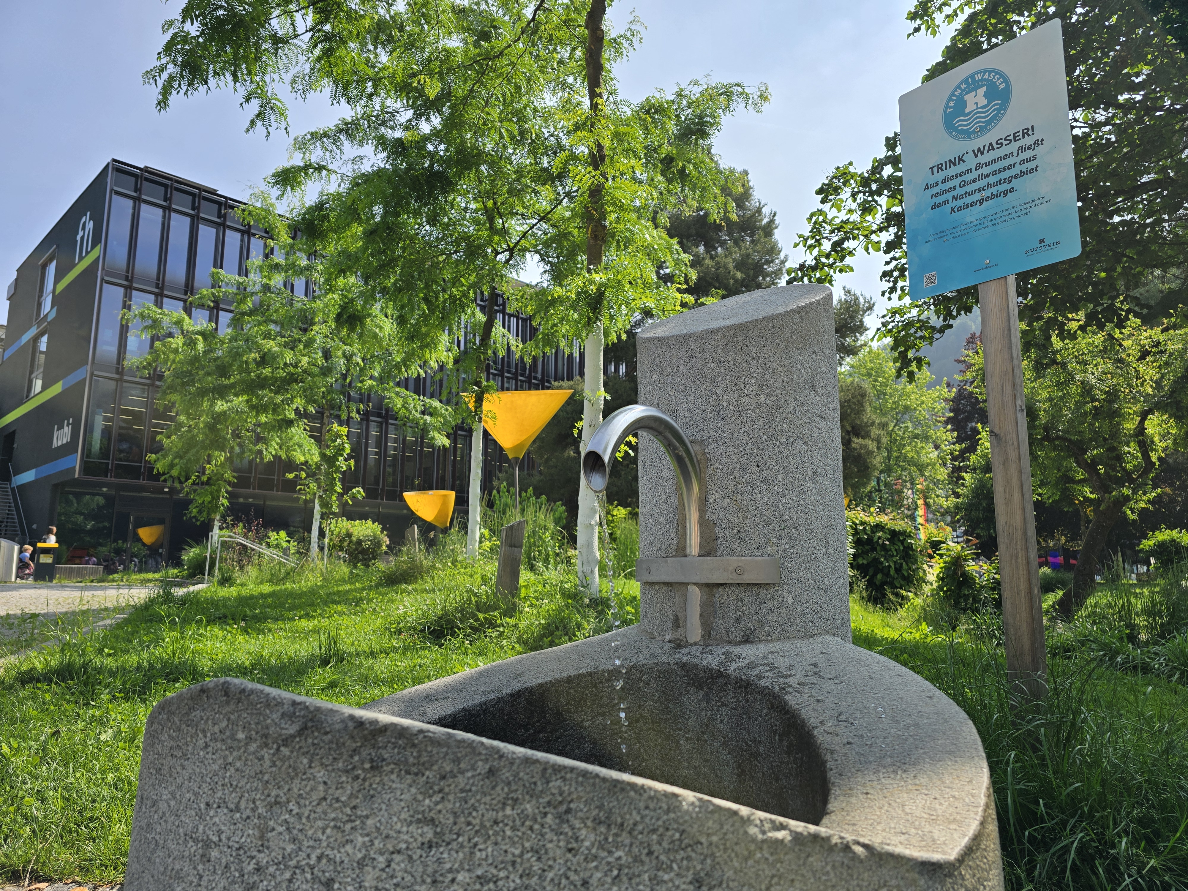 Trinkwasserbrunnen im Stadtpark Kufstein Moderner Trinkwasserbrunnen im Stadtpark Kufstein mit Hinweisschild und FH-Gebäude im Hintergrund.