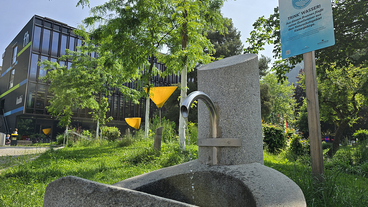 Moderner Trinkwasserbrunnen im Stadtpark Kufstein mit Hinweisschild und FH-Gebäude im Hintergrund.